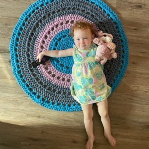 three colored round crochet rug on wood background with baby holding a teddy bear and remote laying on the rug