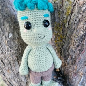 smiling crochet troll doll with blue hair, green skin, brown pants, in a tree background