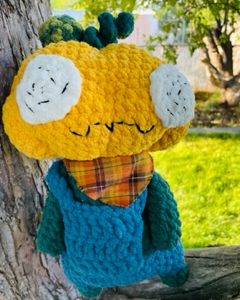 Crochet yellow pumpkin plushie with green body and blue overalls. wearing orange plaid handkerchief around the neck with tree background